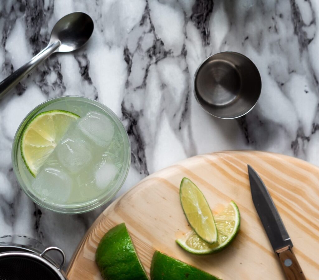 Ein Glas gefüllt mit Eiswürfeln und Limettenscheiben und Flüssigkeit neben einem Holzschneidebrett mit aufgeschnittenen Limettenscheiben und einem kleinen Messer drauf.